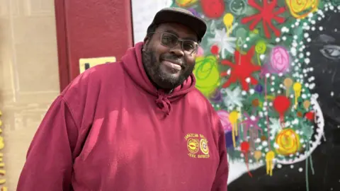 BBC A man wearing a red hooded top with a baseball cap, beard and glasses is stood in front of a boarded up window which has been painted with the image of a lady with bright coloured afro hairstyle 