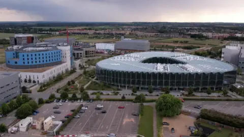 An aerial view of some of the modern buildings at Cambridge Biomedical Campus. There are around six buildings in shot, with a surface car park in the foreground and the horizon in the far distance, beneath a somewhat moody sky.