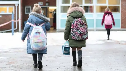 PA Media Two children walking towards a school. They are wearing puffer jackets and fluffy colourful backpacks