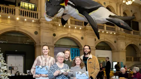 Bristol City Council A group of people hold the book 'Is That Doris?' while stood underneath a model of Doris the pliosaur.