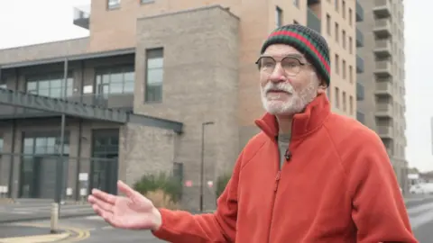 Andrew Boff seen wearing a red jacket and a woollen hat stands on a street, gesturing with one hand, with modern apartment buildings behind him.