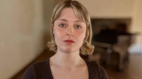 Lizzie Blissett looks directly at the camera. She is wearing a black scoop-neck dress and has her blonde hair in a bob. Behind her, blurred, is a piano.