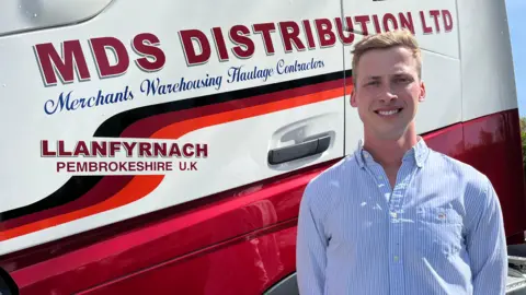 A blonde man in a blue shirt stands in front of the cab of a lorry, branded with the logo of MDS Distribution Ltd.