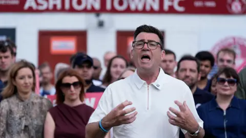 PA Media Andy Burnham, who has black hair gelled into a peak and wearing a white polo-neck t-shirt, speaks in front of a crowd of Labour activists at a non-league football stadium. In the background a red sign reading Ashton Town FC is visible. 