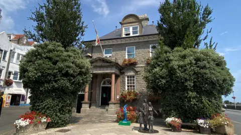 BBC A picture of the outside of the proposed Victor Hugo Centre in Guernsey. It is a large brown building with a statue and flag outside.