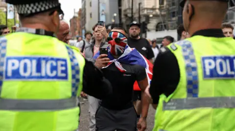 PA Media Two police officers watch as a man in a Union Jack mask films them with a mobile phone