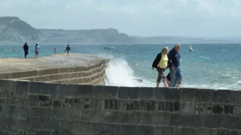 Chris Allen Five people walk along The Cobb as a wave sends up a high plume of spray. The curved breakwater is built from mortared stone