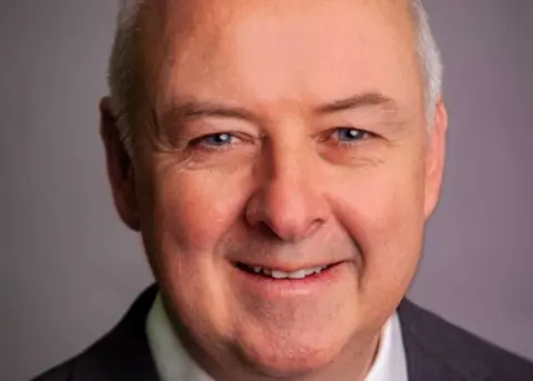 Staffordshire Commissioner Commissioner Ben Adams is looking at the camera, smiling. He is wearing a dark suit and is in front of a purple background.