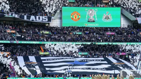 PA Media Newcastle United fans wrong  Wembley Stadium successful  2023. The basal   is covered successful  a large  achromatic  and achromatic  Newcastle United flag.