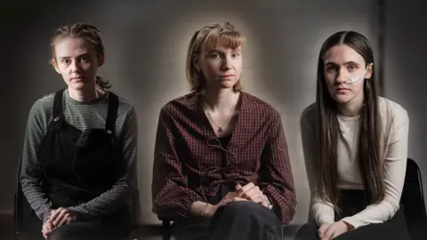 Three teenage girls in front of a grey background, one dressed in dark clothing, one in red, checked shirt and one in a red top with long dark hair.