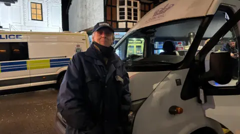A man with grey hair is wearing a navy cap that says "Stafford street pastors" in white writing. He is wearing a matching navy jacket with the same writing on the breast. He is leaning against a white van