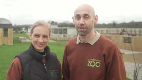 A blonde-haired woman in a black gilet stands next to a man in a brown "Holmes Chapel Zoo" branded jumper. Behind them are wooden enclosures.