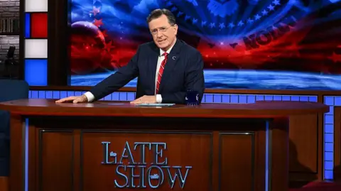 Stephen Colbert sitting behind his desk in The Late Show studio