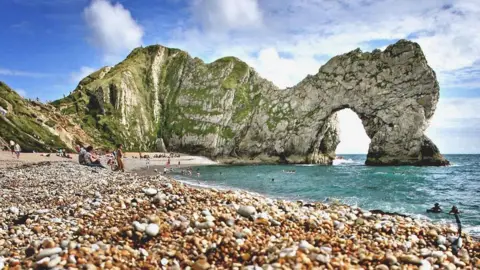 PA Low-angle view from a pebble-covered beach looking toward a dramatic coastal rock arch rising from the sea. The foreground is filled with multicoloured pebbles in sharp detail, leading to turquoise water and a curved shoreline where visitors are scattered along the beach and swimming near the shore. In the background, steep green-and-white cliffs form a natural limestone arch over the water. A bright blue sky with scattered white clouds stretches above the rugged coastline, creating a striking seaside landscape.