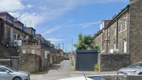 View of a narrow alleyway between rows of stone terraced houses in Bradford, with parked cars on either side of the entrance.