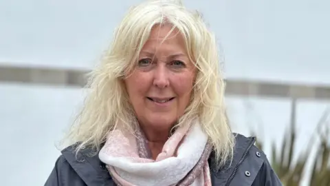 BBC Jane Stevenson, who has long blonde hair and is wearing a dark grey raincoat a pink and white scarf.