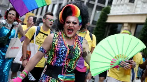 Parade-goers take part in the Pride In London 2026 parade on July 4, 2026 in London, England. Parade goer dressed in black wig with rainbow quiff, multi-coloured leopard print dress and a big green fan, mouth open as if to say 'yay' at the camera. Behind are several others in the parade.