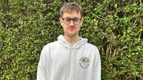 A man stands close infront of a green hedge. He has short brown hair, with the beginnings of a 5 o'clock shadow. He is wearing black glasses with large lenses, and a white hoodie with a round graphic on the left of his chest.