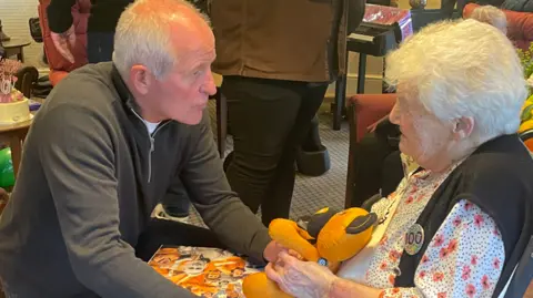 Avery Healthcare Group Steve Bull is kneeling and facing Joan in her chair and holding her hands while he talks to her. Joan is smiling at him and is holding some Wolves memorabilia and is wearing a 100 birthday badge.
