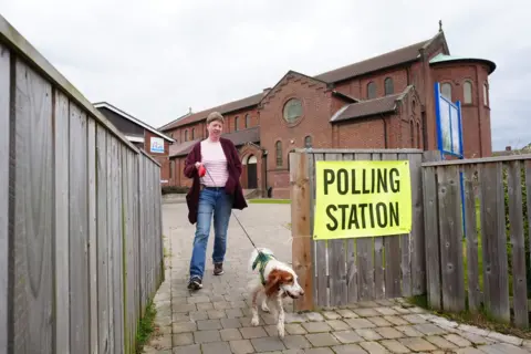 Owen Humphreys / PA Media A person in a pink striped top and blue jeans walks with dog on lead, the dog with white and light brown fur, next to a polling station sign outside the church.