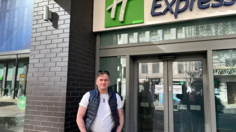 Jonny Humphries/BBC Calumn Donaghey, 30, who has dark blonde hair, a pair of sunglasses on his head and is wearing a dark blue gilet over a white t-shirt. He is standing outside a hotel doorway smiling at the camera