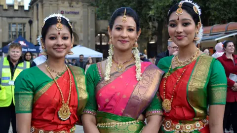 Peterborough Diwali Festival Three women dressed in colourful sarees and gold jewellery with flowers in their hair, smile and look directly at the camera. 