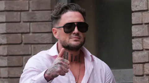 Stephen Bear gives a thumbs up to cameras, wearing an open neck pink shirt revealing a gold chain and neck tattoo