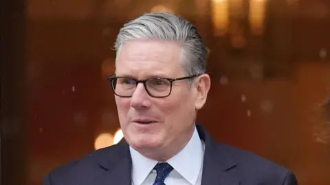 Sir Keir Starmer leaving 10 Downing Street. He has short grey hair, is wearing glasses, a white shirt, blue spotted tie and blue suit. 