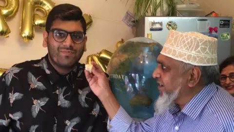 A young man in glasses in black shirt looking at the camera. An elderly man pictured next to him feeding him birthday cake. The man is wearing a blue striped shirt and a beige and white Islamic hat