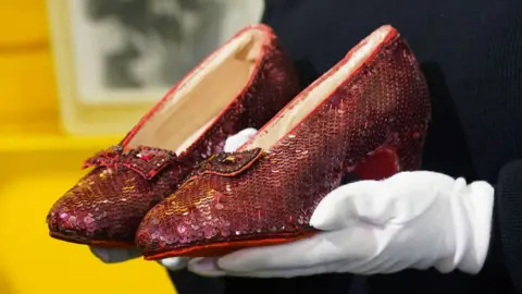 A close up view of a pair of famous Ruby slippers from the movie 'The Wizard of Oz' held by hands wearing white gloves.