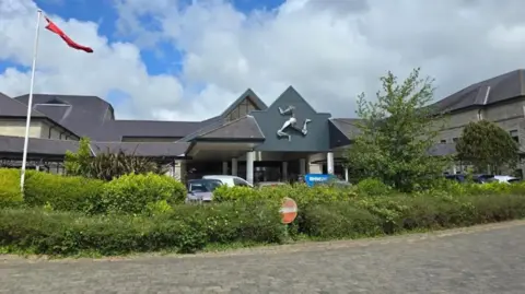 BBC The entrance to Nobles Hospital in Douglas. There is a green hedgerow, and on the left there is a large flagpole flying the Manx national flag. Above the entrance, a large silver triskelion emblem is displayed.