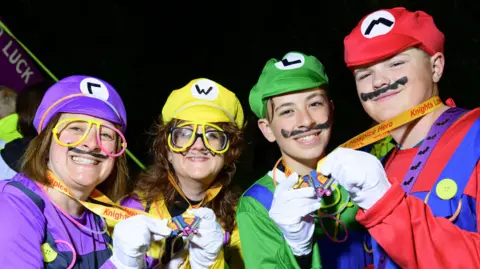 Nick Butcher Photography/St Elizabeth Hospice Four people stand together dressed in different coloured Super Mario costumes. They have medals around their necks that they hold in their hands and face toward the camera.