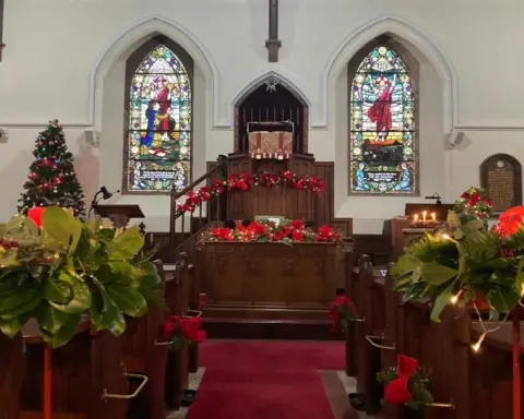 Mitchell family The inside of Applegarth Church from the aisle looking towards the alter, with a row of pews on either side, decorated with greenery and red ribbons. Two stain glass windows are positioned on either side of the alter and there's a Christmas tree in the corner.