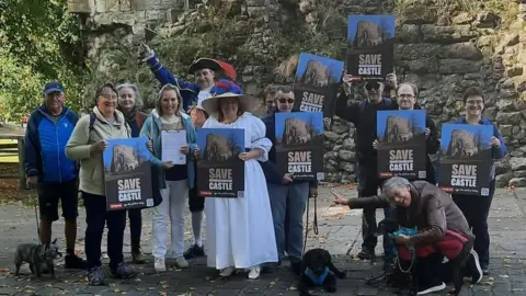A mixed group of people gather outside Knaresborough Castle, holding signs which read 'Save Knaresborough Castle.