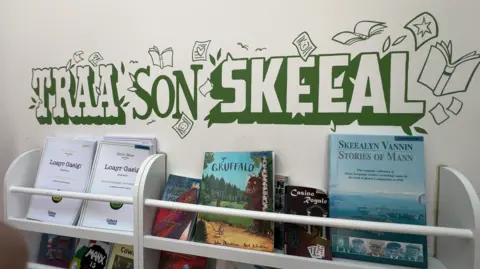 A white shelf holding Manx language books with the words Traa Son Skeeal painted in green on the wall above it.