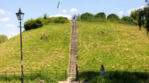 East Herts Council Stone steps up a grass hill leading to a mound with trees and bushes