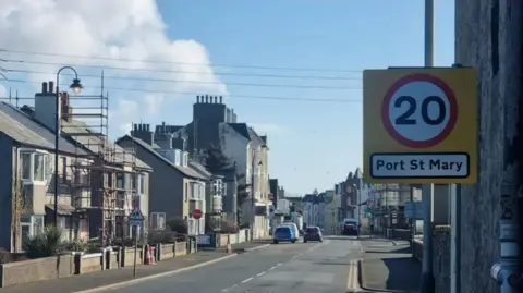 Buildings lining a tarmacked road in Port St Mary village. In the foreground there is a yellow sign with a number 20 in a white circle with a red trim in the middle attached to a pole. It also reads Port St Mary in black writing on a white background.