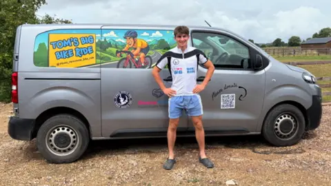 Tom Dunlop in cycling shorts and t-shirt, he is standing in front of a silver van which has artwork of a boy on a bike and reads 'Tom's Big Bike Ride'.