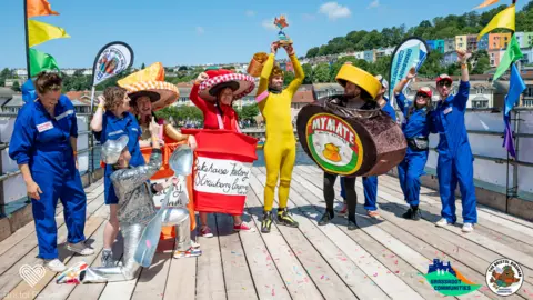 Bristol Picture A group of people wearing different costumes – two people are dressed as jam jars, with gingham lids on their heads, and one man is dressed as a jar of Marmite, except the label reads "My Mate". Another is wearing entirely silver and holding a large spoon. The group is celebreating and a man in a yellow bodysuit in the centre holds up a prize.