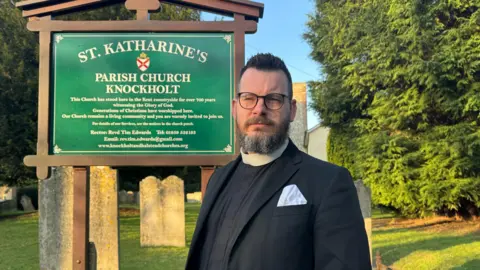 Rev Tim Edwards berdiri di depan tanda gereja untuk St Katherine's in Knockholt. Dia mengenakan gaun klerikal dan memiliki janggut dan kacamata, dengan saputangan di saku atasnya.