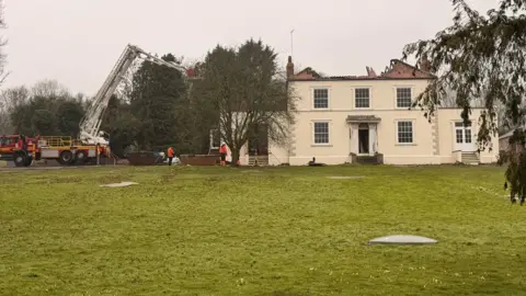 A large detached house with cream-coloured rendered walls. The roof appears to be badly damaged with just a small section to the right of the image being intact. There is a fire engine with an aerial platform to the side.