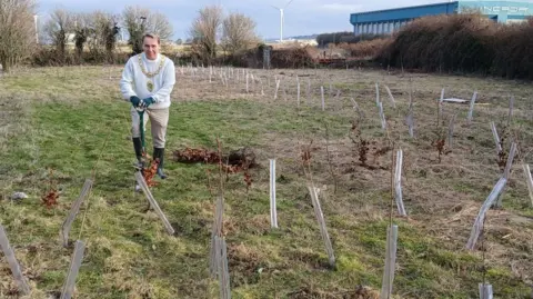 Mayor David Farrar wears a gold chain of office on top of a white jumper and beige trousers while digging a hole in the ground using a spade. He is surrounded by tree saplings protected in plastic tubes on a patch of green land in Siddick. A large blue industrial unit and some wind turbines are in the distance.