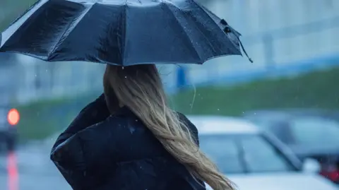 A woman with long hair is pictured in an anonymous, blurred road, holding up an umbrella in torrential rain