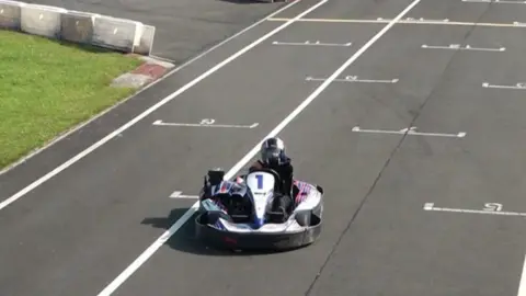 The picture shows a single go‑kart on an outdoor karting track. The kart is driving along a straight section marked with white grid numbers painted on the tarmac. The driver is wearing a helmet and racing gear, and the kart has a large number “1” on its front panel.