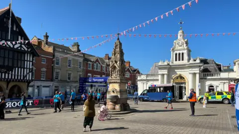 Stuart Woodward/BBC Saffron Walden Square dengan orang -orang yang merenungkan dan air mancur di tengah. Bendera digantung di atas.