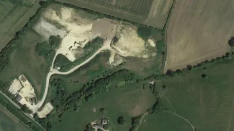 Google Aerial shot of quarry, appearing as white shapes surrounded by green fields. There is a small area of white including buildings - the entrance - at the bottom left of the image, and a road heads up the picture, splitting into two and heading to a larger area of white - the quarry.