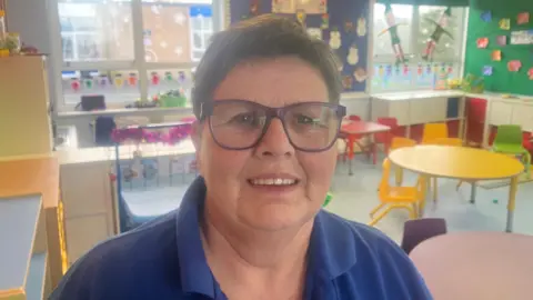 Karen is wearing a blue polo shirt and glasses. She has short brown hair. She is standing in a busy classroom. 