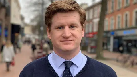 A man with ginger hair wearing a shirt, tie and jersey stands in a street looking into the camera.