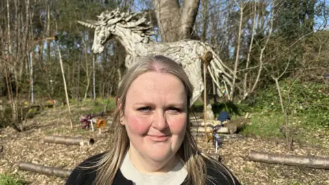 Ben Parker/BBC Debbie Streatfield, with blonde hair and a dark top with a cream top under it. She is standing in front of a large model horse, which is set amongst trees.
