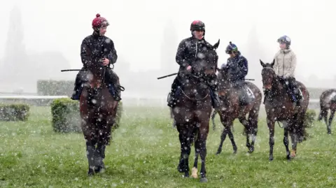 Caballos de medios de PA caminando en la nieve en el hipódromo de Cheltenham con jinetes sentados en la parte superior